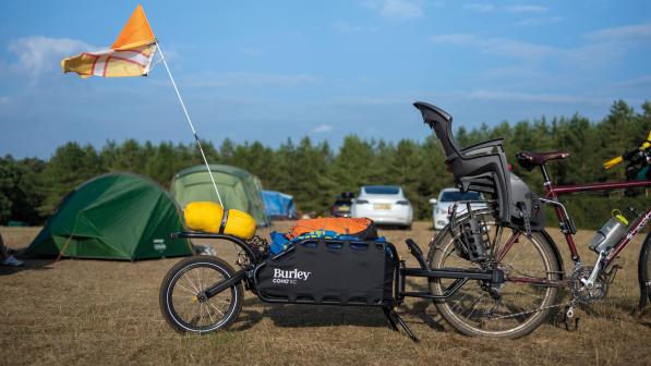 The Coho XC trailer attached to a touring bike with a child seat on the back, packed with stuff, at a campsite with tents and cars in the background