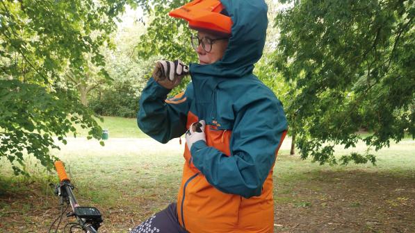 A woman on a mountain bike wearing a blue and orange waterproof jacket with a hood over her orange cycling helmet. She is holding an adjuster to tighten the hood.