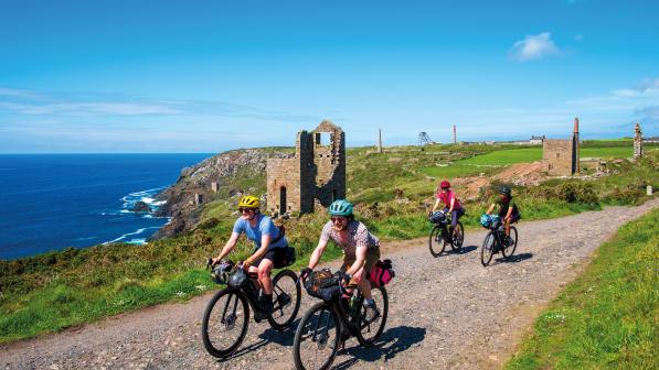 A group of four people are riding along a gravel path with the ocean and mine ruins in the background. They are all riding loaded gravel bikes.