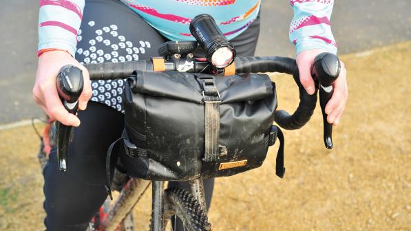 The Restrap Utility Hip Pack, a black waterproof bag, is attached to the handlebar of a drop-bar bike. A person in cycling kit is standing astride the bike with hands on the hoods