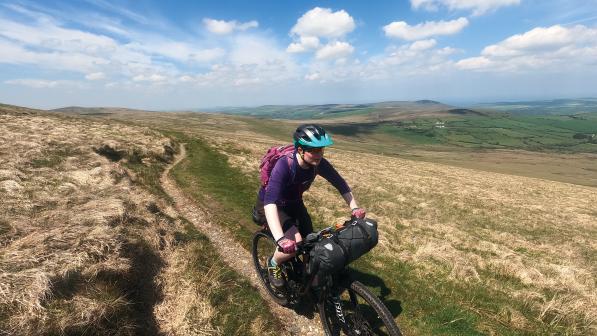A woman is cycling a loaded mountain bike on a bridleway on a high ridge. She is wearing mountain bike gear and a helmet. It's a sunny day.