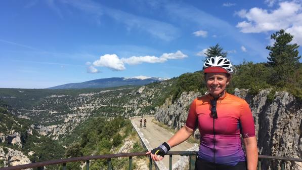 A woman is standing next to a metal fence with one hand on the top. She is wearing a bright, stripy cycling jersey and black cycling shorts. She has on a cycling helmet and gloves and has sunglasses tucked in her jersey. In the background is a mountain road with two cyclists on it. She's smiling at the camera