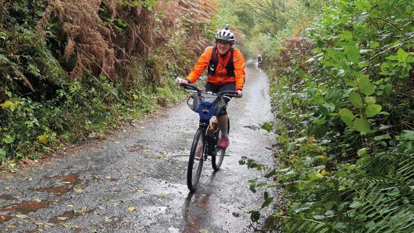 A woman is cycling up a hill on a mountain bike in the rain. She is cycling on a wet, muddy off-road path. She is wearing an orange waterproof jacket, three-quarter-length cycling tights, pink shoes and a cycling helmet.