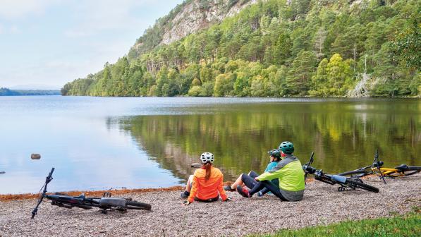 Three people are sitting on the ground on the shore of a loch. They're all wearing cycling kit and there are three bikes lying on the ground next to them. There are pine-clad mountains in the distance