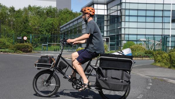 A man in grey T-shirt, navy shorts and orange cycle helmet is riding a black electric cargo bike with a full load on both the front and rear racks. He's on a roundabout in an urban setting