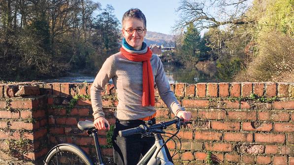 A woman is standing holding a grey hybrid bike. She's outside and is wearing a sweater and jeans with a scarf around her neck. She's standing next to a brick wall over which you can see trees and a river
