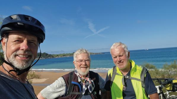Three men with white hair and beards taking a selfie in front of a beach and very blue sea and sky. They're wearing cycling kit and you can just see a bike behind them
