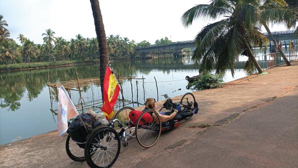 A woman is in a handcycle with a wheelchair attached to the back. She's on a concrete surface with a pond and palm tress in the background