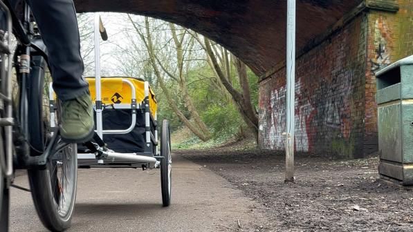 A person is riding a bike pulling a cargo trailer behind, on a tarmac path under a brick bridge. You can only see the back wheel of the bike and the person's left leg and foot