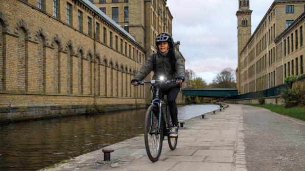 A woman is riding a hybrid bike along a canal towpath in an urban setting. She is wearing warm clothing and a helmet. There is a bright light on the handlebar of the bike
