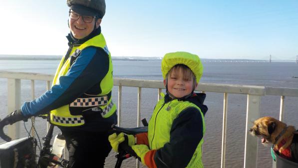 A man and a child are standing astride a tandem bike on a ferry with the river in the background. They are wearing hi-vis kit and cycling helmets. They are smiling at the camera. A border terrier is on the back of the bike
