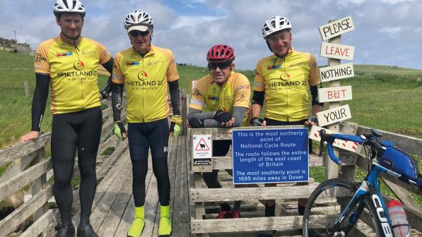 Four men in yellow and black cycling kit, all wearing helmets, are standing on a wooden bridge next to a sign saying they've reached the most northerly point of the National Cycle Route. One bike can be seen in the foreground.