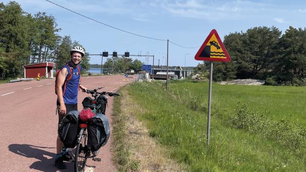 A man is standing by the side of the road holding a packed touring bike. He's wearing shorts and T-shirt and a cycling helmet. He's standing next to a warning sign showing a car falling off a cliff into water.