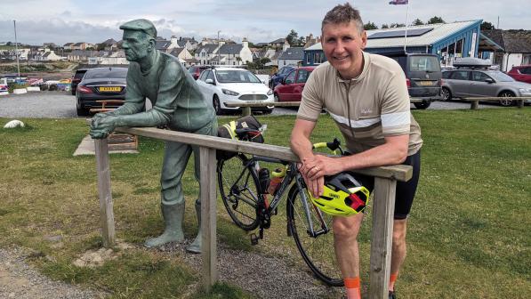 A man is leaning against a wooden fence with his forearms resting on the fence, next to a statue of a fisherman in the same pose. He's wearing cycling kit and there's a bike between him and the statue