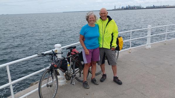 A man and a woman both wearing cycling kit are standing on a promenade with bikes behind them leaning on a metal fence. The sea is in the background