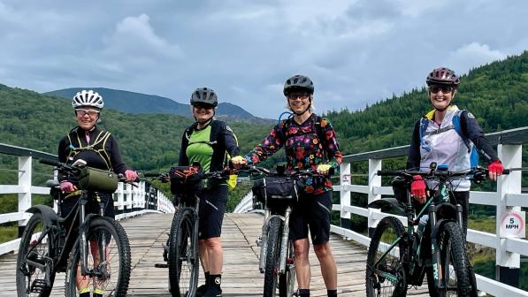 Four women, all with loaded mountain bikes and wearing mountain bike kit and cycle helmets, are standing on a wooden bridge with mountains in the background