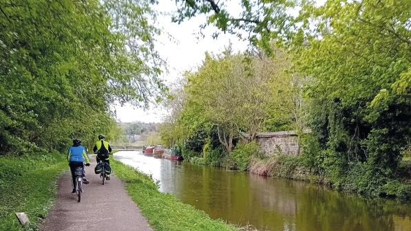 Two people are riding along a paved towpath along the left-hand side of a canal. There are lots of trees and greenery lining the canal. There are three narrow boats in the distance