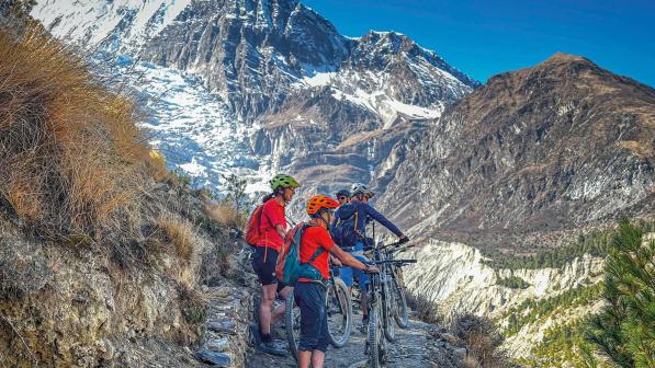 Four people with loaded mountain bikes and wearing mountain bike kit are standing on a stony path looking out over a green valley and snow-covered mountains