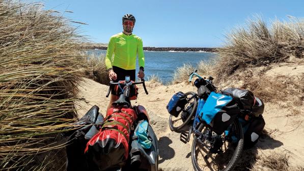 A man is standing with two loaded touring bikes in sand dunes with the ocean in the background. He's wearing cycling kit and helmet