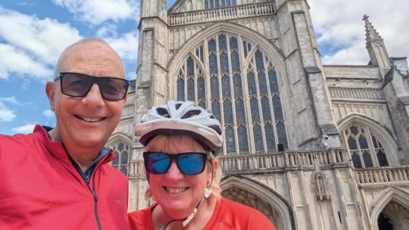 Two people in red tops, both wearing sunglasses and one wearing a cycle helmet, are standing in front of a Gothic-style cathedral