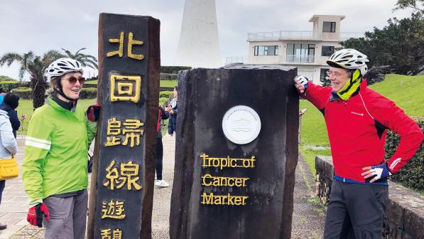 Two people in cycling kit are standing either side of a sculpture of two large rectagonal blocks. One has Taiwanese script on it, the other says 'Tropic of Cancer marker'