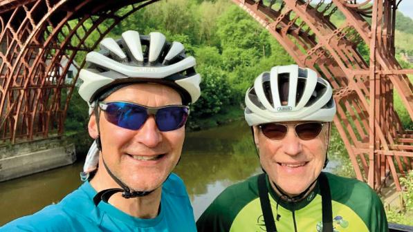 Two men in cycle kit, helmets and sun glasses are posing in front of an iron bridge across a river