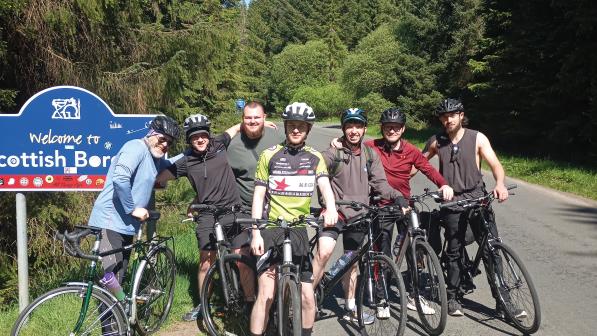 A group of men on bikes are standing to the side of a pine tree-lined road next to a sign reading 'Welcome to the Scottish Borders'
