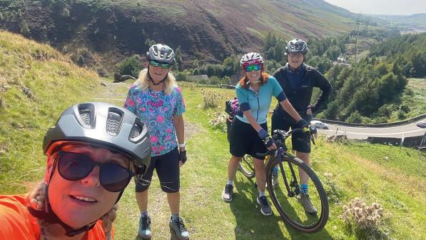 A group of women wearing cycling kit and helmets and one of them holding a mountain bike is standing on a grassy trail in mountains