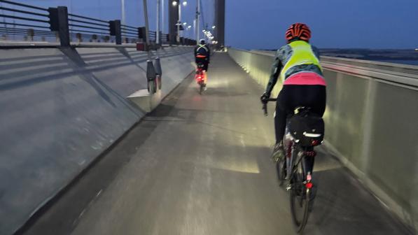 Two people are cycling across a suspension bridge in the early morning. They're wearing cycling it and you can see their lights illuminating the path in front of them