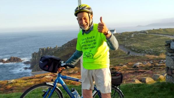 A man stands with a bike under the Land’s End signpost. He is wearing a bright yellow T-shirt and giving a thumbs up to the camera