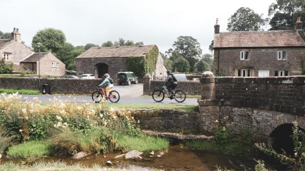 Two people on e-bikes are cycling along a village lane with a stream running alongside them. They have just cycled off a stone bridge. There are stone houses in the background 