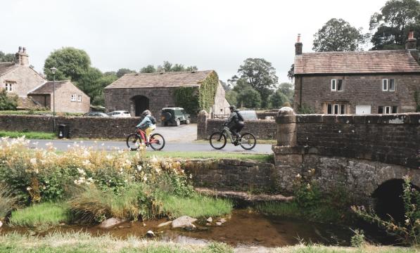 Two people on e-bikes are cycling along a village lane with a stream running alongside them. They have just cycled off a stone bridge. There are stone houses in the background 