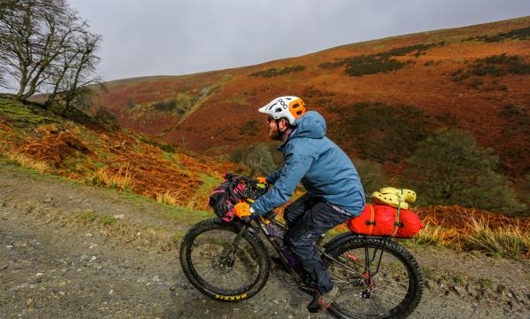 A man rides a mountain bike on a hilly gravel path, his bike weighed down by bikepacking bags. He is wearing full waterproofs and a helmet
