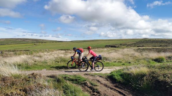 Two people are riding along an off-road path in beautiful countryside. They are riding loaded touring bikes and wearing summer kit.