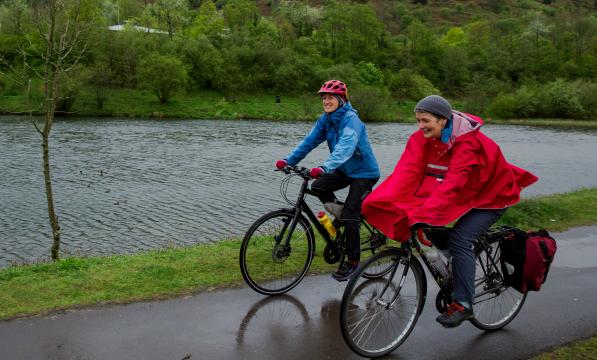 Two women are cycling side by side on a wet path in the rain. One is wearing a blue waterproof, the other a red rain cape. They are both wearing waterproof trousers