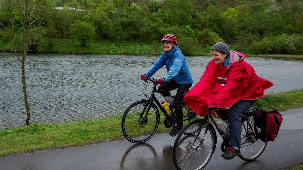 Two women are cycling side by side on a wet path in the rain. One is wearing a blue waterproof, the other a red rain cape. They are both wearing waterproof trousers