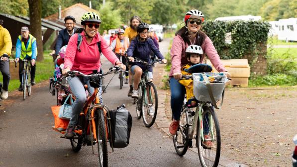 A group of people are cycling on a paved path. Two women are at the front. One is on an orange cargo bike with loaded panniers, the other a hybrid with a basket on the front and a child on a seat on the top bar.