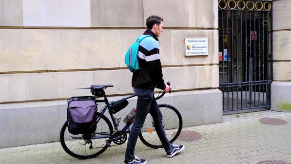A young man walks along a pavement with his bicycle outside a building boasting a plaque that reads 'Clarence Court, Department for Infrastructure, 10-18 Adelaide Street'