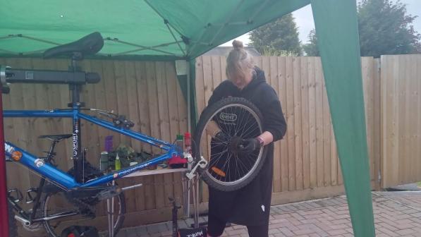 A woman in black clothes and apron is holding a bicycle wheel to put it back on a blue bicycle that's in a stand