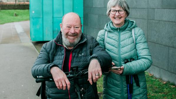 Two people smile at the camera outdoors beside a grey building; one is seated on a cycle and the other stands next to them holding a phone.