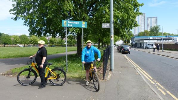 Two men dressed in normal clothes, with cycling helmets, are standing astride yellow electric bikes on a pavement between a road and park