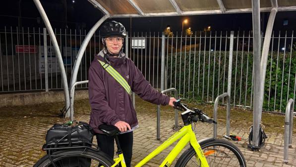 A woman in a purple jacket, black trousers and cycle helmet is standing holding a yellow electric bike with a packed pannier on the rear rack in cycle storage area at night