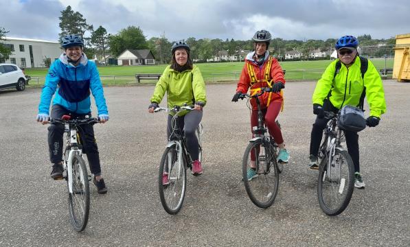 Four people are lined up on their bikes about to set off on a ride. They are wearing brightly coloured jackets and are smiling at the camera.