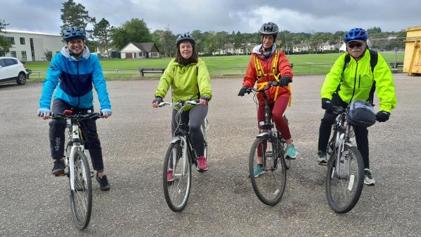 Four people are lined up on their bikes about to set off on a ride. They are wearing brightly coloured jackets and are smiling at the camera.