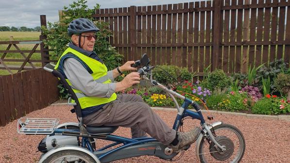 A man wearing cargo trousers and a grey shirt with a yellow hi-vis vest over it and a cycle helmet is sitting on a blue trike in a garden, with a flower bed and wooden fence behind him