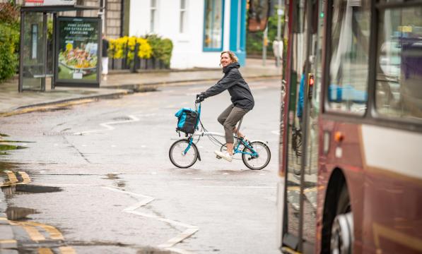 A woman cycles across a road at a pelican crossing in front of a bus. She is riding a Brompton fold-up bike, and is wearing a raincoat and a big smile as she looks at the camera