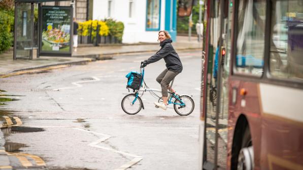 A woman cycles across a road at a pelican crossing in front of a bus. She is riding a Brompton fold-up bike, and is wearing a raincoat and a big smile as she looks at the camera