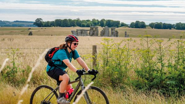 A female cyclists rides past a field in summer with Stonehenge in the background 