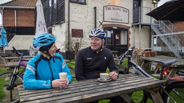 Two cyclists sit at a wooden table in a pub garden, they are holding coffees and chatting