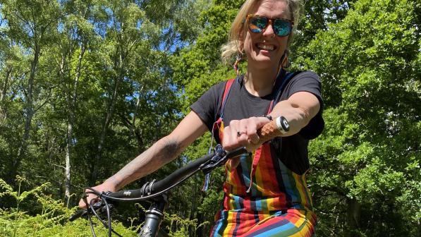 A woman is sitting on a mountain bike on an off-road trail. She's wearing rainbow dungarees and a black T-shirt